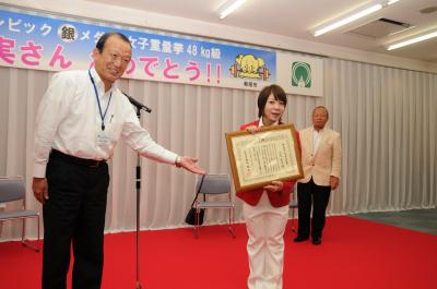 三宅宏実選手が、須田健治市長から贈呈された市民栄誉賞を会場の皆さんに見せている場面の写真です。
